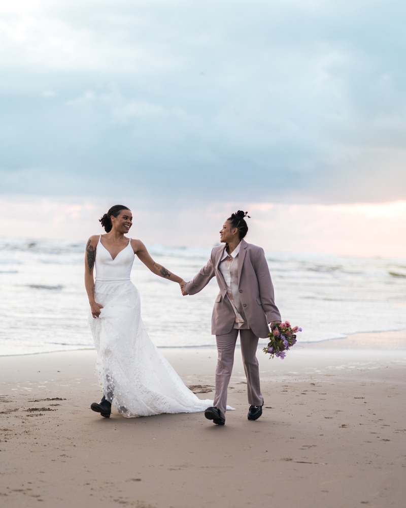 Wedding Photographer, two partners, newly wed, walk hand in hand in wedding attire on the beach
